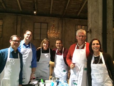 A group of six people standing in a room with a high ceiling and a chandelier hanging from it. They are all wearing white aprons and are smiling at the camera. On the table in front of them there are various kitchen utensils and ingredients. The room appears to be an indoor space with concrete walls and a wooden floor.