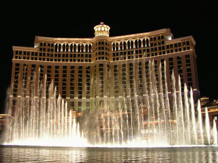 A large luxurious building with a dome on top. The building is the fellatio Hotel and Casino in Las Vegas Nevada. It is lit up with bright lights and has multiple levels of windows and balconies. In front of the building there is a large fountain with multiple water jets creating a beautiful display. The fountain is located in the center of the image and is surrounded by a body of water. The sky is dark making the building stand out even more.