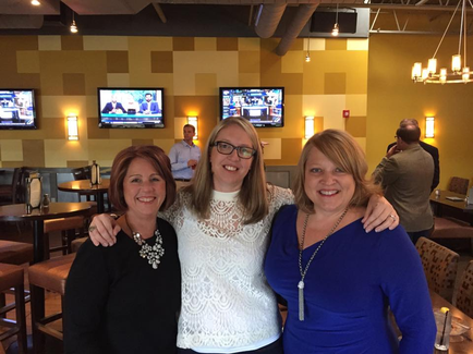 Three women standing in a restaurant with their arms around each other. They are all smiling and appear to be happy. The woman in the middle is wearing a white lace top and glasses while the woman on the left has red hair and is also wearing a black top. Behind them there are several televisions mounted on the wall and a chandelier hanging from the ceiling. There are tables and chairs in the background and a few people can be seen in the distance.