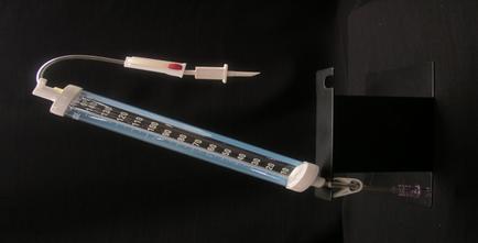 A syringe with a needle attached to it. The syringe is made of plastic and has a long thin tube with a white cap. The needle has a red tip and a white handle. The body of the syringe has a blue and white striped pattern with numbers on it indicating the amount of liquid inside. On the right side of the image there is a black metal stand with a small purple light bulb attached to the bottom right corner. The stand appears to be made of metal and is placed on a black surface.