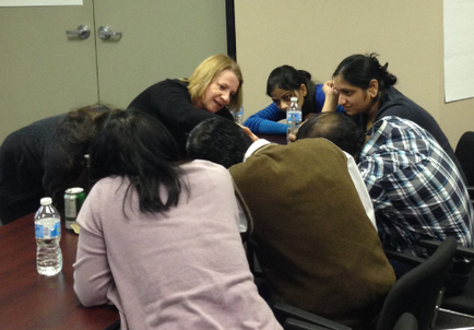 A group of six people sitting around a wooden table in a meeting room. There are six people in total three women and three men all of whom appear to be engaged in a conversation. The woman in the center of the image is leaning over the table with her head in her hands while the other three women are leaning over her shoulder. They are all looking at each other and seem to be in deep thought. There is a water bottle on the table in front of them. The room has a door and a whiteboard on the wall in the background.