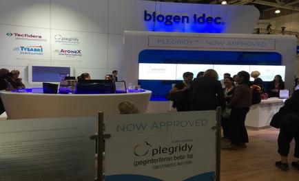 A trade show booth with a blue and white color scheme. The booth has a sign that reads "Biogen idea" and "pleurisy - Now Approved". There are several people gathered around the booth some of them are looking at the sign and others are engaged in conversation. On the left side of the image there is a desk with a computer monitor and other items on it. In the background there are several other booths with logos of different companies. The overall atmosphere of the booth appears to be busy and professional.