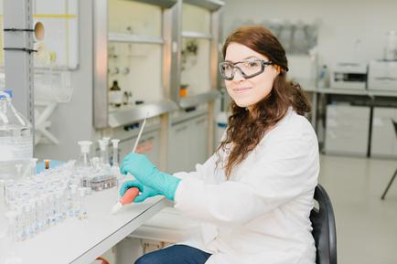 A young woman in a laboratory setting. She is wearing a white lab coat blue gloves and safety goggles. She has long brown hair and is sitting on a chair in front of a white countertype with multiple test tubes and flasks on it. The woman is holding a pipette and appears to be conducting a scientific experiment. In the background there are shelves with various laboratory equipment and shelves with bottles and other scientific equipment. The overall atmosphere of the image is focused and professional.