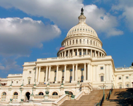 A photograph of the United States Capitol building in Washington DC. The building is a large neoclassical structure with a dome-shaped dome on top and a statue of a man on a pedestal on the right side. The dome is white with columns and arches and the American flag is flying on the left side of the dome. The sky is blue with a few clouds scattered across it. In front of the building there is a set of stairs leading up to the entrance. The image is taken from a low angle looking up at the building.