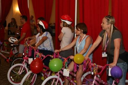A group of young women standing in front of a row of pink bicycles. The bicycles are decorated with colorful balloons and streamers. The women are wearing red helmets and appear to be participating in a party or event. The background is a red curtain and there are other people in the background. The overall mood of the image is festive and cheerful.