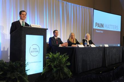 A panel discussion taking place on a stage. There are four people sitting at a long table with nameplates in front of them. The table is covered with a black tablecloth and there are plants on either side of the table. On the left side there is a man standing behind a podium with a microphone and a nameplate that reads "PAIN MATTERS". On the right side two women and a man are sitting at the table smiling and looking at the camera. In the background there are two large screens with the words "Thank You" and "Pain Matters" displayed on them.