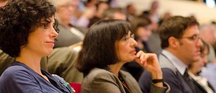 A group of people sitting in a lecture hall attentively listening to a presentation. The focus of the image is on a woman with curly hair who appears to be in deep thought. She is wearing a blue blouse and has a badge on her lapel. Next to her there is another woman with shoulder-length dark hair also wearing a gray blazer. She has her hand on her chin and is looking off to the side with a serious expression on her face. Behind her a man in a suit and tie is also listening attentively. The background is blurred but it seems to be a large room with other people in the audience.