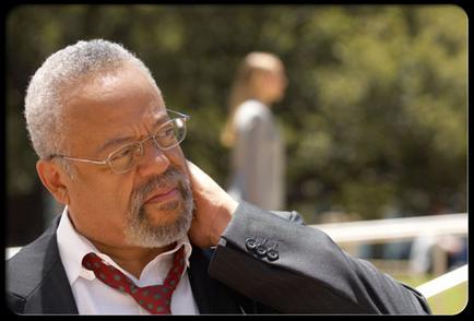 A close-up portrait of an African-American man. He is wearing a suit and a red tie with white polka dots. He has a pair of glasses on and is looking off to the side with a thoughtful expression on his face. The background is blurred but it appears to be an outdoor setting with trees and a person walking in the distance.