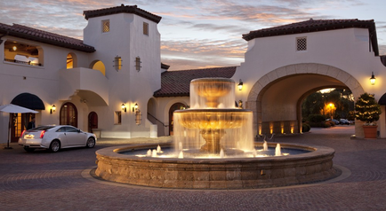 A beautiful courtyard with a large fountain in the center. The fountain is made of stone and has multiple tiers of water cascading down from the top. The water is illuminated by warm lights creating a peaceful and serene atmosphere. The courtyard is surrounded by a white building with arches and pillars and there are several potted plants and trees in the background. A white car is parked in the courtyard and the sky is a beautiful orange and pink hue indicating that it is either sunrise or sunset. The overall mood of the image is peaceful and inviting.