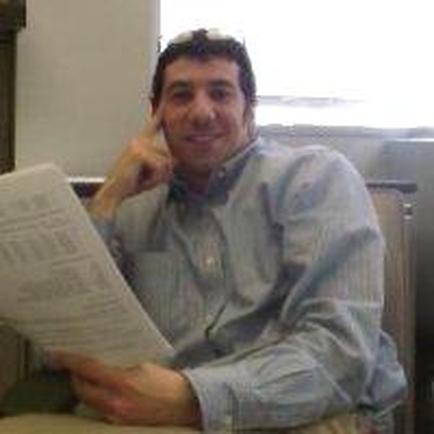 A man sitting on a couch and reading a newspaper. He is wearing a light blue collared shirt and has a pair of sunglasses on his head. He has a smile on his face and is resting his chin on his hand as if he is deep in thought. The newspaper he is reading is open and he is holding it in his left hand. The background appears to be a room with a window and a bookshelf.