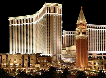 A night view of the Venetian Hotel and Casino in Las Vegas Nevada. The hotel is a tall white building with a curved facade and multiple levels. It is lit up with bright lights creating a beautiful contrast against the dark night sky. In front of the hotel there is a large clock tower with a golden dome and intricate details. The clock tower is surrounded by palm trees and other buildings and there are several other buildings in the background. The overall atmosphere of the image is luxurious and opulent.