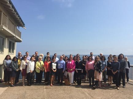 A group of people standing on a balcony overlooking the ocean. There are around 20 people in the group all of them are smiling and posing for the photo. They are standing in front of a building with a balcony on the left side of the image. The sky is blue and the ocean can be seen in the background. The group appears to be of different ages and ethnicity's and they are all dressed in casual clothing. Some of them have sunglasses on suggesting that they are taking a photo or group photo.