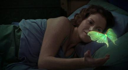 A young woman lying in bed with her head resting on a blue pillow. She is wearing a blue tank top and has red hair. The woman is smiling and looking at the camera. In her hand she is holding a green butterfly that is glowing in the dark. The background is dark and the overall mood of the image is peaceful and serene.