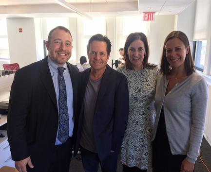 Four people standing in an office space with an exit sign in the background. On the left is a man wearing a black suit and tie on the right is a woman wearing a gray cardigan and a black and white patterned dress and in the middle is a middle-aged man with dark hair and a beard. All four people are smiling and posing for the photo. They appear to be happy and relaxed.