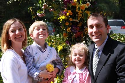 A family of four posing for a photo in front of a beautiful floral arrangement. The family consists of a mother father and two young children. The mother is on the left side of the image wearing a white blouse and a pearl necklace. The father is in the center with the mother on the right side and the two children on either side of him. The children are all smiling and looking at the camera. The background is filled with a variety of colorful flowers and greenery including yellow pink purple and orange. There is a white car parked in the background. The overall mood of the photo is happy and joyful.