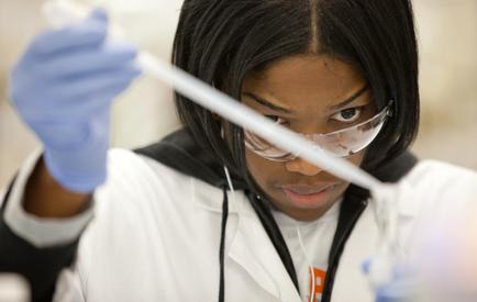 A young African-American woman wearing a white lab coat and blue gloves. She is holding a pipette in her right hand and appears to be working in a laboratory. She has a pair of safety goggles on her face and is looking intently at the pipette. The background is blurred but it seems like she is focused on her work.