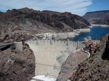 A large dam located in a mountainous area. The dam is made of concrete and has multiple levels with a large body of water on the right side. The water is a deep blue color and appears to be calm. On the left side of the dam there are several large structures including a large building with a dome-shaped structure on top which is likely a hydroelectric dam. There are also several smaller structures scattered around the dam. The sky is blue and there are power lines running across the image. The mountains in the background are steep and rocky and the overall landscape is barren and barren.