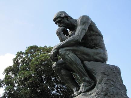 A photograph of a statue of a man sitting on a rock. The statue is made of bronze and appears to be of a muscular man with his head resting on his hand. He is sitting with his legs crossed and his arms resting on the rock. His body is hunched over and his head is tilted downwards as if deep in thought. The man's facial expression is one of contemplation or deep thought. In the background there is a tree and a clear blue sky. The overall mood of the image is somber and contemplative.