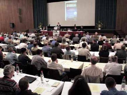 A large conference room with a stage in the center. The room is filled with people sitting at long tables with white tablecloths and water bottles on them. The people are facing the stage where a man is giving a presentation. The stage is covered with a red tablecloth and there is a projector screen at the front of the room. The walls are made of wood and there are two large windows on either side of the stage. The audience is seated in rows of chairs facing the audience and attentively listening to the presentation.