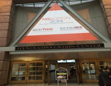 The entrance of the haynes Convention Center in Boston Massachusetts. The building has a large triangular sign with the words "boston 2014 actress" written in red and white. The sign is attached to the top of the building with a metal frame. Below the sign there is a sign that reads "haynes Convention Centre" in black letters. The entrance has two large glass doors and a brick wall on the left side. There are a few people walking in front of the entrance.