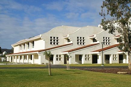 A large two-story building with a white exterior and a curved roof. The building has multiple windows on the upper level and a red tiled roof on the lower level. The front of the building has a large lawn with a few trees and shrubs. The sky is blue and there are a few clouds in the background. There is a street lamp on the right side of the image and a few other buildings visible in the distance.