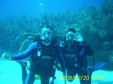 Two scuba divers posing for a photo in the ocean. They are both wearing full scuba gear including oxygen tanks swim fins and goggles. The diver on the left is wearing a blue wetsuit with the word "tills" written on it while the diver in the middle is wearing an oxygen tank. They both have their arms stretched out in front of them as if they are posing for the photo. The background is filled with coral reefs and other marine life. The water is clear and blue and the date "2008/02/20 15:37" is visible in the bottom right corner of the image.