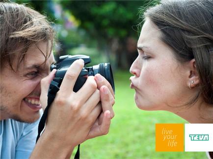 A man and a woman taking a photo together with a DSLR camera. The man is holding the camera in his hands and the woman is kissing him on the cheek. They are standing in a park or garden with trees and grass in the background. The woman has her eyes closed and the man is looking at her with a smile on his face. The image appears to be taken during the day.