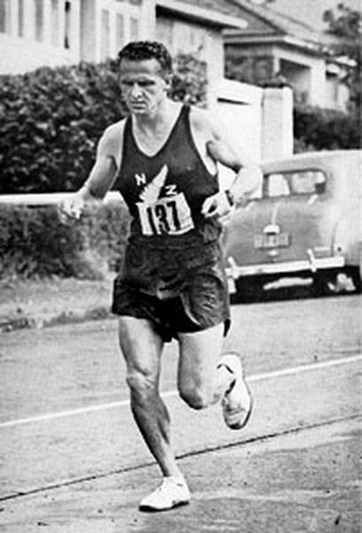 A black and white photograph of a man running on a street. He is wearing a sleeveless tank top with the number "137" on it and black shorts. He appears to be in the middle of a race as he is running with determination and focus. The man is running on the right side of the image with his arms and legs pumping as he runs. In the background there is a car parked on the street and a building on the left side. The photograph appears to have been taken in the early 20th century.