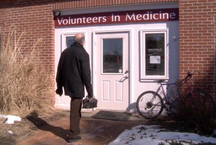 An elderly man walking towards the entrance of a brick building with a sign that reads "Volunteers in Medicine." The man is wearing a black coat and carrying a black bag in his left hand. He is walking towards a white door with a glass window on the right side. There is a bicycle leaning against the door and a doormat in front of the door. The ground is covered in snow and there are some bushes and shrubs on the left side of the image.