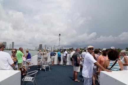 A group of people standing on the deck of a boat looking out at the city skyline. The sky is cloudy and the water is calm with a few boats visible in the distance. The people are dressed in casual summer clothes and some are wearing hats and sunglasses. They appear to be engaged in conversation and enjoying the view. In the background there are tall buildings and skyscrapers suggesting that the photo was taken in a city. The overall mood of the image is relaxed and casual.