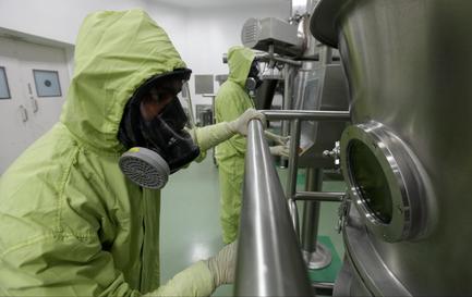 Two people wearing green protective suits and masks in a laboratory setting. The person on the left is wearing a full-body suit with a hood and a gas mask covering their face. They are standing in front of a large stainless steel tank with a round window on the right side. The tank appears to be part of a larger machine or equipment. The background shows other machines and equipment in the laboratory. The people are working on the tank and appear to be in the process of cleaning or disinfecting it.