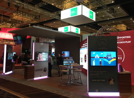 A trade show booth with a large screen on the right side of the booth. The booth is decorated with a red and white color scheme and has a sign that reads "Supporting each play". There are several people in the booth some standing and some sitting at tables. On the left side there is a large TV screen with a picture of a woman on it. In the background there are several other booths with different designs and colors. The overall atmosphere of the image is modern and professional.