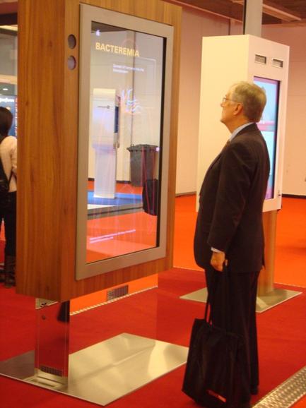 A man in a suit standing in front of a large screen with the word "bacteria" written on it. The screen is mounted on a wooden stand and is placed on a red carpeted floor. The man is holding a black bag in his left hand and appears to be looking at the screen intently. In the background there are other people and a white wall.