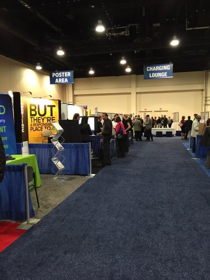 A large exhibition hall with a blue carpeted floor and white walls. There are several booths on the left side of the image each with a sign that reads "But they're not allowed". On the right side there is a charging lounge sign. People are gathered around the booths some standing and some walking around. The hall appears to be well-lit with bright lights.