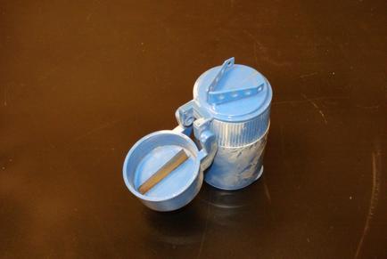A small blue tin canister with a lid and a handle. The canister is made of metal and has a rusted and weathered appearance. The lid has a small handle on top and a small opening on the side. The handle is attached to the lid with a metal ring. The body of the canister has a textured surface with a pattern of small circles and lines. There is also a small wooden stick sticking out of the lid. The background is a dark brown surface.