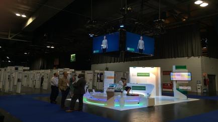 A large exhibition hall with a blue carpeted floor and white walls. There are several large screens hanging from the ceiling displaying images of people. On the left side of the image there are rows of white booths with green and white signs. In the center of the hall there is a white booth with a desk and a computer monitor. A group of people are gathered around the booth looking at the screens. The overall atmosphere of the exhibition hall is modern and professional.