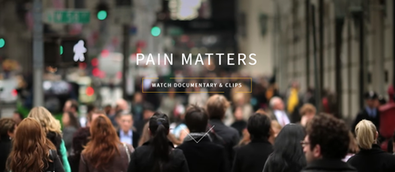 A busy street scene with a crowd of people walking on the sidewalk. The street is lined with buildings on both sides and there are traffic lights and street signs visible in the background. The people in the crowd appear to be of different ages and genders and they are all dressed in casual clothing. In the center of the image there is a text overlay that reads "Pain Matters: Watch Documentary & Clips".