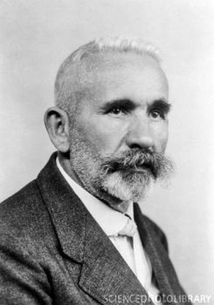 A black and white portrait of a man. He appears to be in his late 60s or early 70s with a bald head and a full beard. He has a serious expression on his face and is looking directly at the camera. He is wearing a suit and tie and his hair is neatly combed back. The background is plain and blurred putting the focus on the man's face.