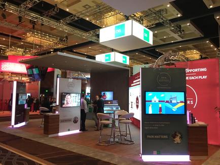 A trade show booth with a modern design. The booth is located in a large hall with a high ceiling and a chandelier hanging from it. The walls are decorated with red and white banners and there are several large screens on the walls. On the right side of the booth there is a large screen displaying a video game with the words "Supporting each play" and "Pain Matters" above it. There are several people standing around the booth and interacting with the game. In the background there are other booths and tables with various items on display. The overall atmosphere of the image is lively and vibrant.