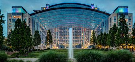 A large modern building with a curved glass facade. The building is surrounded by trees and greenery and there is a fountain in front of it. The sky is dark and the building is lit up with colorful lights. The overall atmosphere of the image is peaceful and serene.