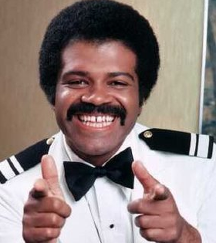 A close-up portrait of a man in a formal attire. He is wearing a white shirt with black stripes and a black bow tie. He has a big smile on his face and is making a thumbs up gesture with both hands. His hair is styled in an afro and he appears to be in his late 40s or early 50s. The background is blurred but it seems to be a room with a wooden paneled wall.