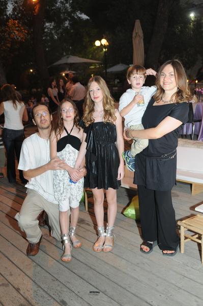 A family of five posing for a photo on a wooden deck at night. There are four people in the photo two adults and three children. The adults are standing close together with the adults on the left side of the image and the children on the right side. The adults are smiling and appear to be happy. The children are standing in front of them with one of them holding a young boy in their arms. The boy is wearing a white shirt and blue jeans while the adults are wearing casual clothes. The background shows a crowd of people tables and umbrellas suggesting that the photo was taken at an outdoor event or gathering. The deck is lit up with string lights creating a warm and cozy atmosphere.