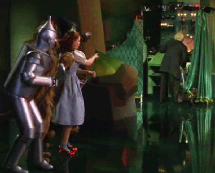 A scene from a movie or TV show. On the left side of the image there is a man dressed as a knight in full armor wearing a helmet and holding a sword. He is standing on a stage with a green curtain in the background. In front of him there are two women one wearing a white dress and the other wearing a black dress both of whom appear to be dancing. The woman in the white dress is holding a large green object possibly a piece of paper or fabric and is reaching out to touch it. The man in the black suit is standing behind the woman in white and he is looking down at her with a serious expression on his face. The stage is lit with bright lights and there are various props and equipment scattered around.
