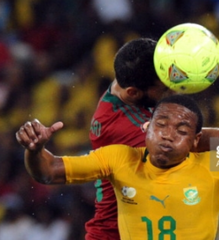 Two soccer players in action during a match. The player on the left is wearing a yellow jersey with the number 18 on it and is in possession of the ball. He is jumping up to head the ball with his right arm while the player in the red jersey is attempting to tackle him. The background is blurred but it appears to be a stadium with spectators in the stands. Both players are focused on the ball and appear to be in the midst of a battle for possession.