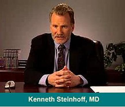 A screenshot from a video interview with Kenneth Steinhoff MD. He is sitting at a desk with his hands clasped together in front of him. He has a serious expression on his face and is wearing a dark suit and tie. Behind him there is a bookshelf and a plant. The background is a plain white wall.