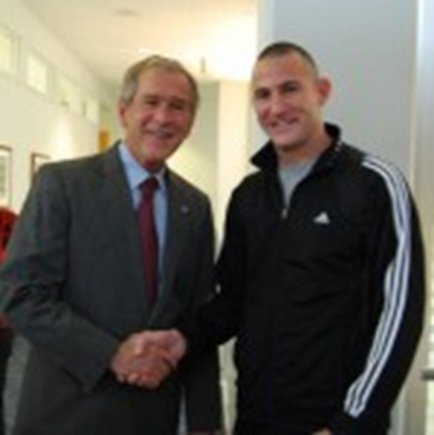 Two men shaking hands in a hallway. The man on the left is older and is wearing a dark suit and a red tie. He has a smile on his face and appears to be in a good mood. On the right is younger and is also smiling. He is also wearing a black jacket with white stripes on the sleeves. The hallway has white walls and there are framed pictures hanging on the walls.