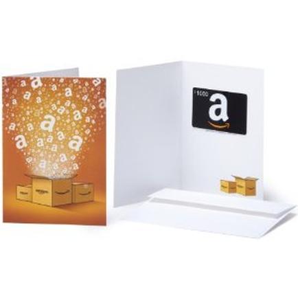 A white greeting card with an orange background. On the left side of the card there is an illustration of a stack of cardboard boxes with the letters "a" and "b" on them. The boxes are arranged in a way that they appear to be floating in the air. The card is placed on a white surface and on the right side there are three small cardboard boxes stacked on top of each other. The background is white and the card has a black Amazon logo on the top right corner.