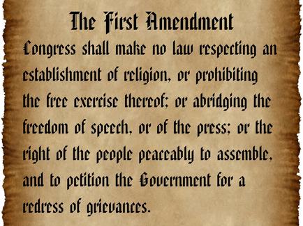 A text-based graphic that reads "The First Amendment: Congress shall make no law respecting an establishment of religion or prohibiting the free exercise thereof or abridging the freedom of speech or of the press; or the right of the people peacefully to assemble and to petition the Government for a redress of grievances." The text is written in black font on a beige-colored background. The text appears to be old and worn with some creases and wrinkles on the edges.