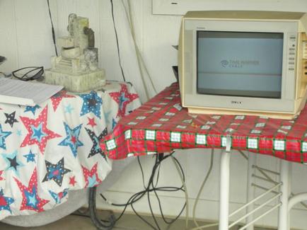 An old-fashioned television set on a white metal stand with a red and green plaid tablecloth. The television is turned on and the screen is turned off. On the left side of the image there is a table covered with a white tablecloth with blue and red stars on it. Next to the table there are some papers and a small statue of a bird. The background is a white wall with a window.