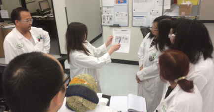 A group of six people in a laboratory setting. They are all wearing white lab coats and are gathered around a table with papers and books on it. The person in the center of the image is holding a piece of paper and appears to be explaining something to the group. To his left there is a man wearing a white lab coat and glasses who seems to be a scientist or a researcher. Behind him there are several other people including a woman with long dark hair and a man with short dark hair who are also wearing lab coats. The group is looking at the paper and seems engaged in a discussion. The room is well-lit with natural light coming in from the windows in the background.